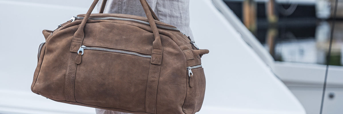 The Riomar Club Travel Bag in brown leather, carried at the marina with a yacht behind